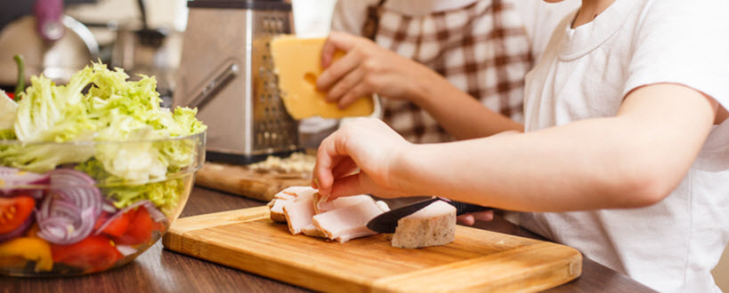 Boy cutting pieces of meat while a girl grates a brick of cheese in the background.